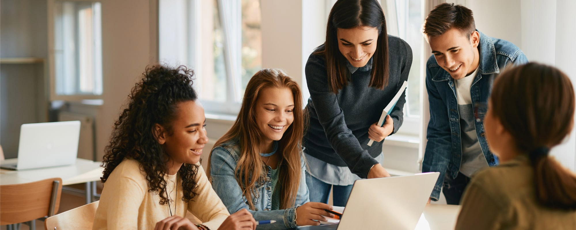 Un groupe de jeunes adultes souriants et collaborant autour d'un ordinateur portable dans une salle de classe lumineuse.